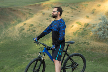 Obraz premium Cyclist in shorts and jersey on a modern carbon hardtail bike with an air suspension fork standing on a cliff against the background of fresh green spring forest