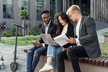 Confident international co workers sitting on wooden bench in front of office and discuss results of joint work.