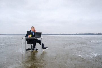 elderly bearded smiling businessman in suit and skates, smiling while talking on smartphone, works with laptop on table in the middle of a frozen lake. Copy space for text, work on vacation concept