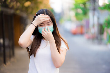 Fototapeta premium Teenage woman in a green medical face mask is coughing, and she touches her forehead with her hands to take body temperature. Prevent the spread of germs.