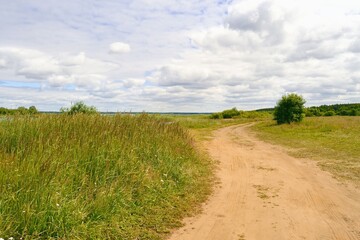 Summer landscape with country road, green meadow grass, beautiful cloudy sky and green forest on the horizon.