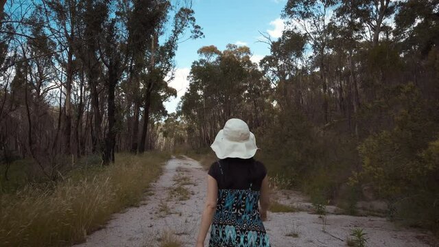 A young Asian girl wearing a decorative dress with a white hat walking down a sandy pathway surrounded by a Gum tree forest, located in the Australian landscape.