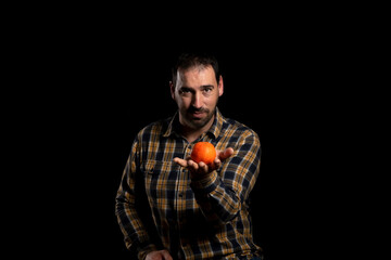 Fototapeta premium Young bearded man holding and offering an orange on the palm of his hand, isolated on black studio background. Copy space