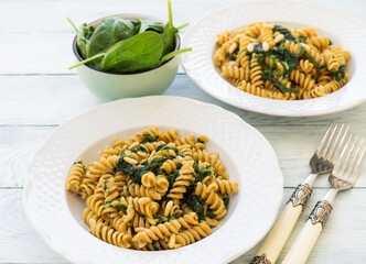 Whole grain fusilli pasta dish with spinach, pine nuts and lactose free cream sauce on white wooden table. Delicious vegan lunch.