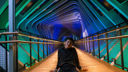 a man who sits casually on a pedestrian bridge at night covered with colorful lights in the Senayan...