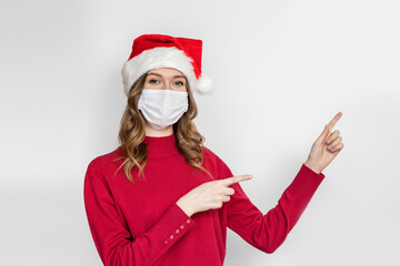 Fototapeta na wymiar Young woman in New Year's holiday hat, red sweater, protective medical mask, points her finger at copy space isolated on white studio background. Coronavirus, New Year's quarantine