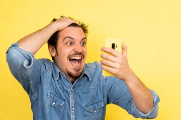 Foto op Canvas A young Caucasian male showing excitement after seeing something on the phone against a yellow background © Adrian Ferrer Iglesias/Wirestock