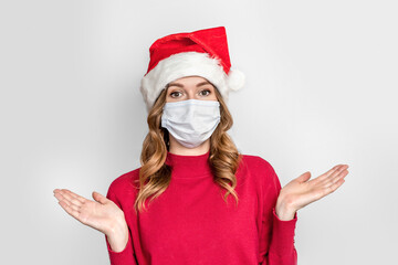 Fototapeta na wymiar Surprised young woman in a New Year's festive hat, red sweater wearing a medical protective face mask spreads her arms to the sides isolated on white studio background. Girl in disbelief
