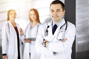 Professional middle aged doctor with a stethoscope and crossed arms at work, standing together with his female colleagues in a sunny clinic. Perfect medical service in a hospital. Medicine and