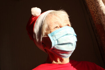Fototapeta premium Senior woman near the window in the sunny morning with a Santa hat wears a medical mask due to the coronavirus.