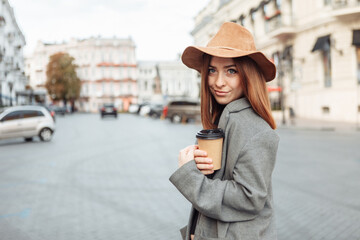 Stylish millennial woman in a slicker coat and felt hat jerks a coffee cup on the go while walking in a european city