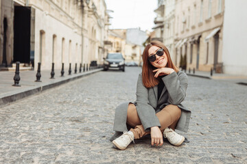 Fototapeta premium Young funny cheerful pretty woman is sitting in the middle of a European city street.