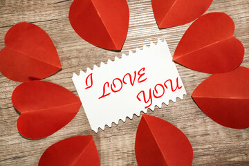 I love you is written on a white sheet that lies on a wooden surface among red hearts.