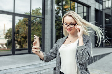 Confident business woman makes selfie on smartphone against the background of business office. Portrait of young attractive blonde girl in business suit and glasses