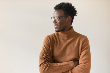 Obraz premium Body language. Horizontal shot of fashionable young African American man with stubble folding arms and looking away with confident happy smile, his look expressing confidence and optimistic attitude