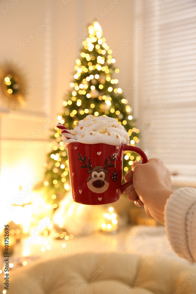 Poster Woman holding cup of delicious drink with whipped cream near Christmas tree indoors, closeup