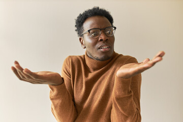 Frustrated young African man in trendy glasses shrugging shoulders in bewilderment, having confused doubtful look, spreading palms, feeling uncertain about making decision, weighing all pros and cons