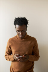 Vertical image of serious focused young black man in stylish turtleneck sweater holding smart...