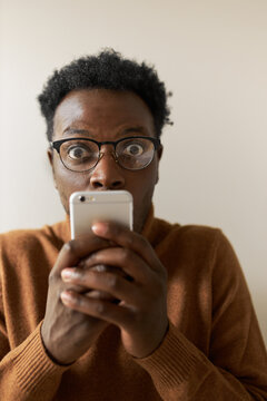 Technology, Internet, Privacy And Confidential Information. Close Up Of Nervous Fearful Young African Male In Spectacles Holding Cell Phone In Both Hands, Widening Eyes, Seeing Something Astonishing