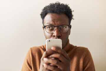 Electronic gadgets and online communication. Studio image of suspicious young dark skinned man typing text message via online messenger on mobile. Thoughtful black guy reading sms, texting reply - obrazy, fototapety, plakaty
