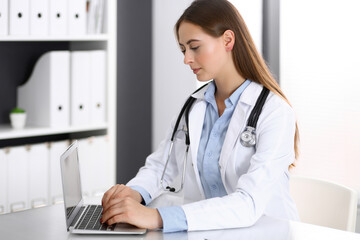 Doctor woman typing on laptop computer while sitting at the desk in hospital office. Physician at work. Medicine and health care concept