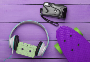 Flat lay hipster composition. Cruiser board, audio cassette, headphones, camera on purple wooden background. Retro 80s entertainments. Summer fun. Top view