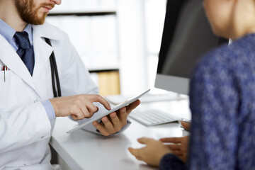 Unknown red-bearded doctor and patient woman discussing current health examination while sitting and using tablet computer in clinic, close-up. Medicine concept