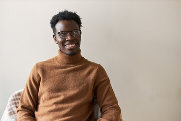 Obraz premium Attractive cheerful young dark skinned unshaven man sitting on chair, waiting for his turn in queue for job interview, smiling joyfully, feeling confident, wearing casual sweater and spectacles