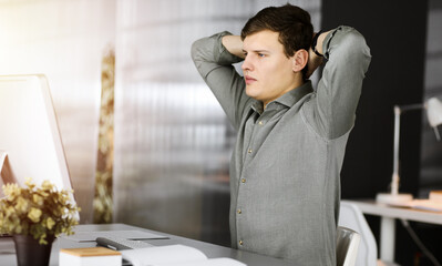 Young businessman with brown curly hair in a green shirt is stretching his hands after working hard on computer in sunny office