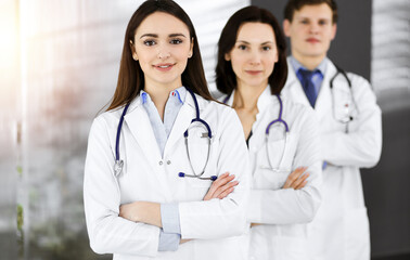 Fototapeta premium Group of young professional doctors standing as a team with arms crossed in sunny hospital. Medical help, insurance in health care, best disease treatment and medicine concept