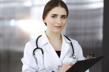 Friendly female doctor standing and holding clipboard in modern clinic. Portrait of cheerful smiling physician