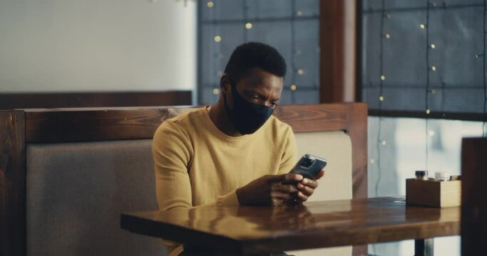 Black guy in mask using smartphone in cafe