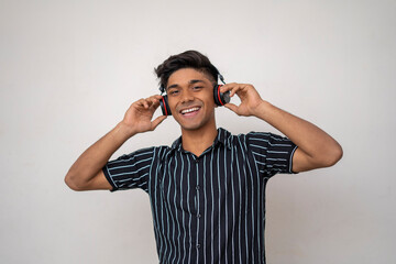 portrait of happy young indian man in half shirt, holding headphone with both hand and smiling listening music.