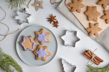 Decorating of sweet savory gingerbread cookies with violet icing served on gray plate cooked for winter holidays on white wooden table with textile, cutters, fir tree branches. Horizontal, flat lay