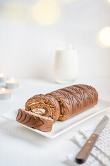 Brown chocolate sponge roll cake with white cream served on plate with knife on textile towel and glass of milk on white wooden table with candles and defocus lights at winter holiday. Vertical