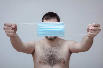 a man with a naked torso holds a medical protective masks in front of his face, covering it to hide...