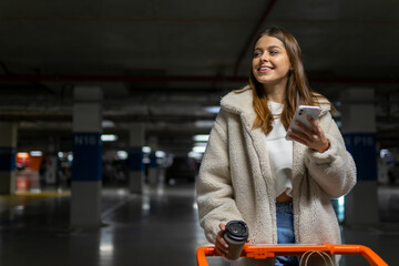 Pretty young woman with smartphone in hands and shopping trolley in the underground parking of shopping mall. Portrait of female shopper with shopping cart