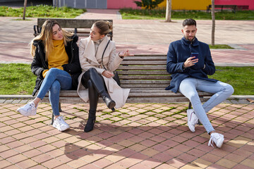 Two beautiful teenagers share secrets, gossip while a boy looks at his cell phone, they are sitting...