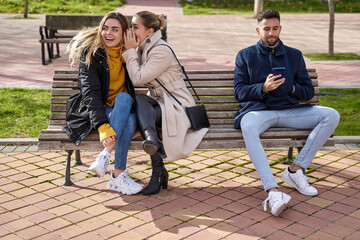 Two beautiful teenagers share secrets, gossip while a boy looks at his cell phone, they are sitting on a park bench.