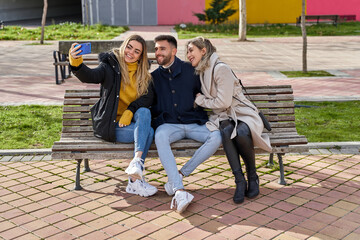 Three young smiling friends sitting on a park bench. Two girls and a boy taking self-portraits on their smartphones. Models posing on the street. Concept of friendship and technology