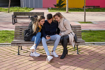 Three young friends sitting outdoors and looking at their cell phones. A group of people sitting on a park bench outdoors and watching a video on their smartphone. Concept of friendship and technology