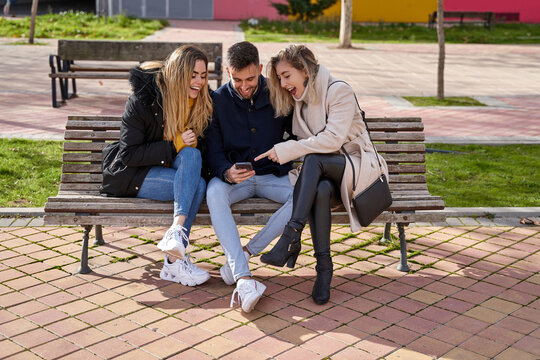 Three young friends sitting outdoors and looking at their cell phones. A group of people sitting on a park bench outdoors and watching a video on their smartphone. Concept of friendship and technology