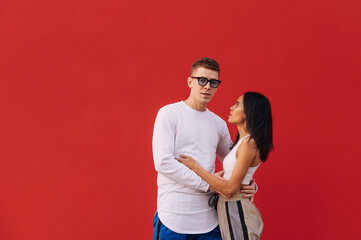 Beautiful stylish couple of young people isolated on red background, embracing, man looking at camera and woman at lover. Man and woman in embracing on a red wall background.Multicultural love.