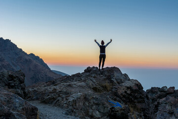 A female tourist standing on the peak of a rocky mountain watching sun rising in Darband valley in autumn in dawn against colorful sky in the Tochal mountain.
