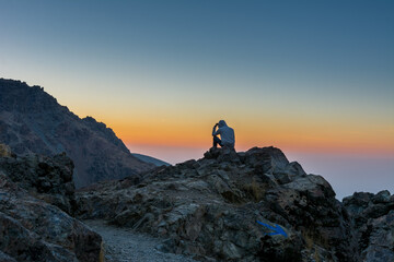 A tourist sitting on the peak of a rocky mountain watching sun rising in Darband valley in autumn in dawn against colorful sky in the Tochal mountain.