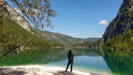 Fototapeta na wymiar A man with a big backpack standing at the shore of the Pragser Wildsee, a lake in South Tyrolean Dolomites. High mountain chains around the lake. The sky and mountains are reflecting in the lake