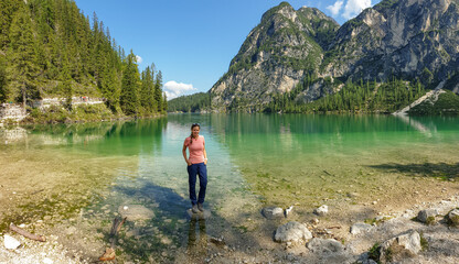 A woman standing on a boulder at the shore of the Pragser Wildsee, a lake in South Tyrolean Dolomites. High mountain chains around the lake. The sky and mountains are reflecting in the lake. Calmness