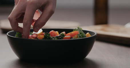 man hand add coppa slices to make salad with kale, and cherry tomatoes in black bowl on concrete countertop