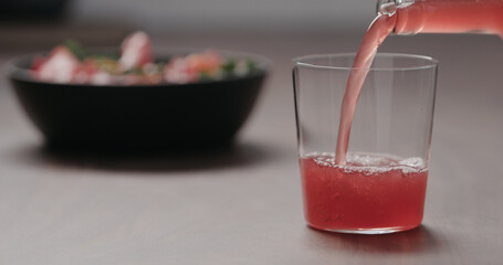 pour pink fizzy drink in a tumbler glass on walnut table with salad on background