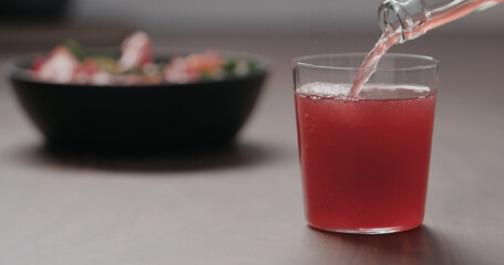 pour pink fizzy drink in a tumbler glass on walnut table with salad on background
